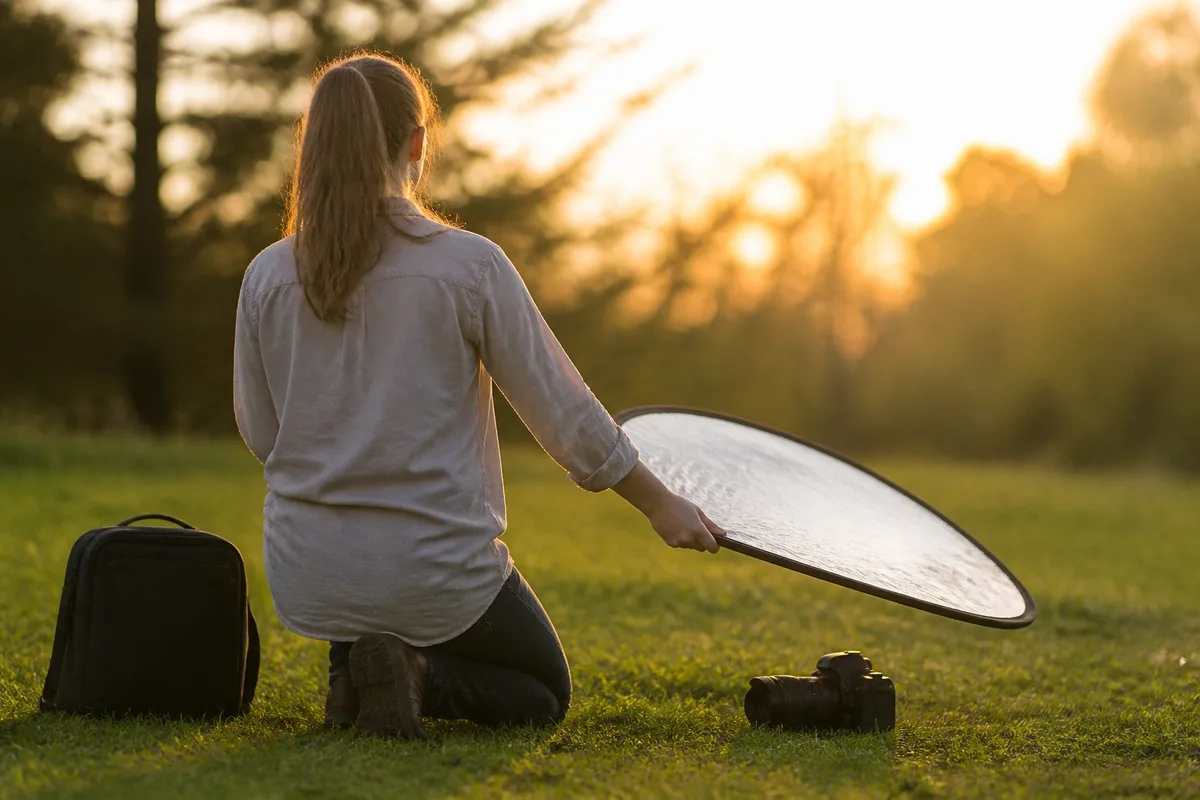 Photographe agenouillée orientant un réflecteur pliant vers un sujet en fin de journée